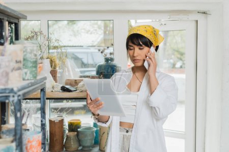 Junge asiatische Künstlerin mit Kopftuch spricht mit Smartphone und digitalem Tablet, während sie im Hintergrund in einer unscharfen Keramik-Werkstatt arbeitet, kreativer Prozess der Töpferei