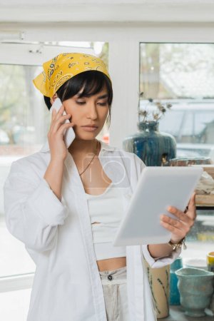 Retrato de joven artista asiática en pañuelo para la cabeza y ropa de trabajo usando tableta digital y hablando en smartphone en taller de cerámica, proceso creativo de fabricación de cerámica