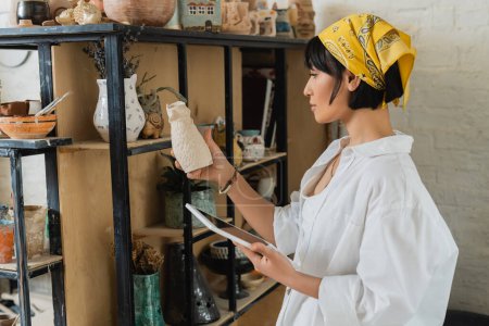 Photo for Side view of young asian female artisan in headscarf and workwear holding digital tablet with blank screen and clay sculpture and standing in ceramic workshop, creative process of pottery making - Royalty Free Image