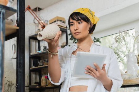Junge asiatische brünette Kunsthandwerkerin in Arbeitskleidung und Kopftuch hält ein digitales Tablet in der Hand und betrachtet die Tonskulptur, während sie im Hintergrund in einer verschwommenen Keramikwerkstatt steht, Prozess der Töpferei