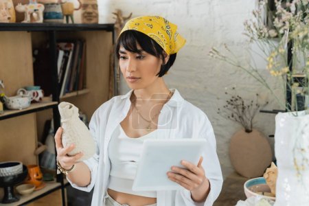 Junge brünette asiatische Kunsthandwerkerin mit Kopftuch und Arbeitskleidung, die Tonskulpturen hält und digitale Tablets benutzt, während sie in der Keramikwerkstatt steht, kreativer Prozess der Töpferei