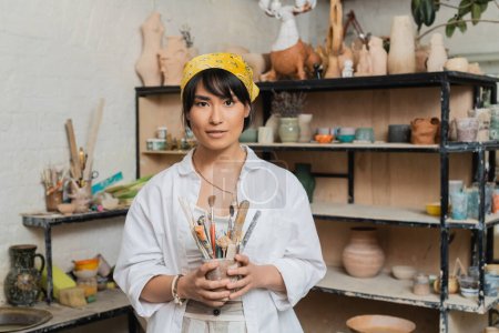 Young brunette asian female artisan in workwear and headscarf holding pottery tools and looking at camera while standing in ceramic workshop, creative process of pottery making