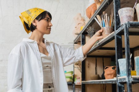 Junge asiatische Künstlerin in Arbeitskleidung und Kopftuch, die Töpferwerkzeuge ins Regal in der Nähe von Tonprodukten stellt und in einer Keramikwerkstatt arbeitet, kreativer Prozess der Töpferei