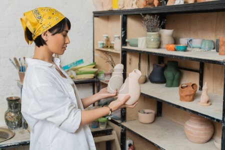 Photo for Side view of young asian female potter in headscarf and workwear holding clay sculptures while standing near rack in ceramic workshop, pottery studio scene with skilled artisan - Royalty Free Image
