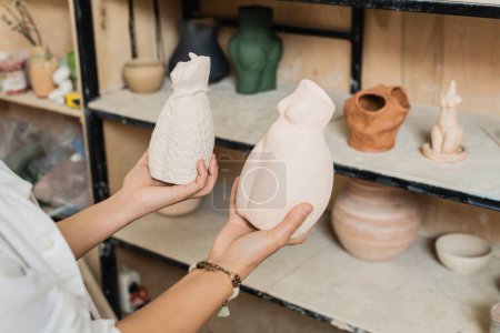 Cropped view of young female artisan in workwear holding clay sculptures while standing near blurred rack in ceramic workshop, pottery studio scene with skilled artisan