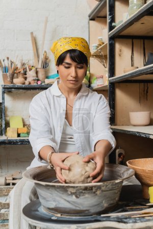 Young brunette asian artisan in headscarf and workwear holding clay while working with pottery wheel near rack and blurred pottery tools in workshop, pottery studio scene with skilled artisan