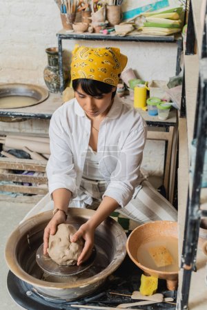 Junge brünette Töpferin mit Kopftuch und Arbeitskleidung, die Ton auf Töpferscheibe hält, während sie neben Schüssel mit Wasser in Keramikwerkstatt sitzt, Töpferatelier-Szene mit geschickter Kunsthandwerkerin