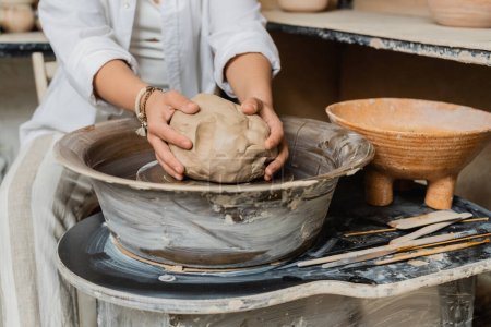 Cropped view of blurred female artisan in workwear putting clay on pottery wheel near tools and bowl in ceramic workshop, pottery studio scene with skilled artisan