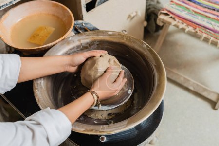 High angle view of young female artisan in workwear molding clay while working with pottery wheel near bowl with water in workshop, pottery studio scene with skilled artisan