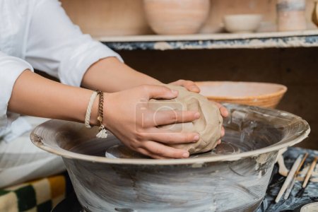Cropped view of young female artisan in workwear putting clay on pottery wheel near tools and bowl in ceramic workshop, pottery studio scene with skilled artisan