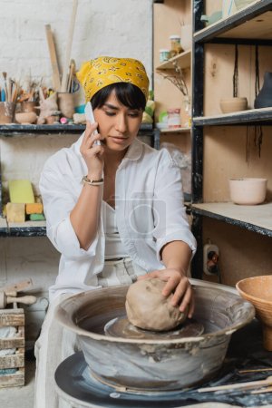 Young asian female artisan in headscarf and workwear talking on smartphone and putting clay on pottery wheel in ceramic workshop, artisan crafting ceramics in studio