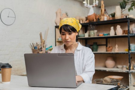 Young asian female artisan in headscarf and workwear using laptop while working near coffee to go in blurred ceramic workshop at background, pottery artist showcasing craft