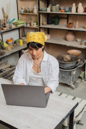 Vista de ángulo alto de la joven mujer asiática artesana en pañuelo para la cabeza y ropa de trabajo usando el ordenador portátil mientras está sentado en la mesa y trabajando en un taller de cerámica borrosa, artista de cerámica mostrando artesanía