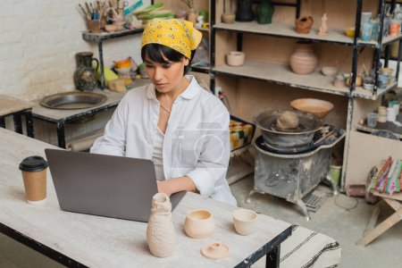 Young brunette asian craftswoman in headscarf and workwear using laptop while working near coffee to go and clay products on table in ceramic workshop, pottery artist showcasing craft