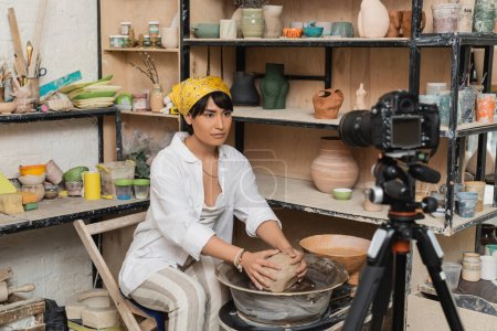 Young asian female artisan in headscarf and workwear putting clay on pottery wheel near digital camera on tripod in ceramic workshop, pottery artist showcasing craft, influencer 