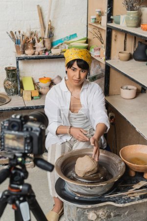 Junge asiatische Künstlerin in Kopftuch und Arbeitskleidung blickt auf Digitalkamera, während sie im Hintergrund neben Ton auf Töpferscheibe in Keramikwerkstatt sitzt, Töpferkünstlerin zeigt Handwerk