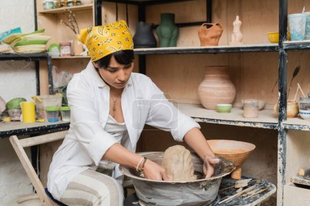 Joven mujer asiática artesana en ropa de trabajo y pañuelo en la cabeza moldeado de arcilla en la rueda de cerámica cerca de esculturas en rack y herramientas en taller de cerámica, artista de cerámica mostrando artesanía