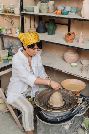 Young asian brunette artisan in headscarf and workwear pouring water on clay on pottery wheel near bowl and rack in ceramic workshop, pottery artist showcasing craft