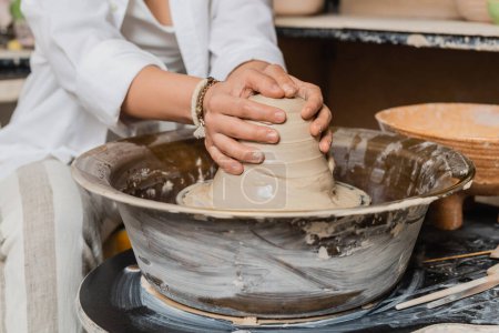 Cropped view of blurred craftswoman in workwear molding wet clay on pottery wheel near bowl at background in ceramic workshop, pottery studio workspace and craft concept