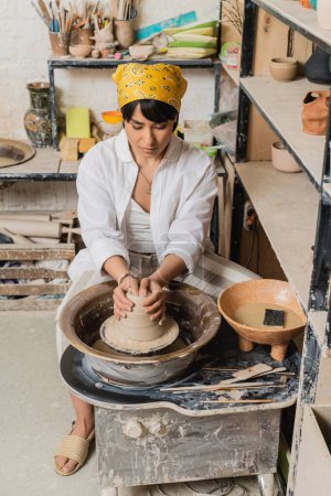 Young asian female artisan in headscarf in workwear molding clay on pottery wheel near bowl with water and sponge near rack in ceramic workshop, pottery studio workspace and craft concept