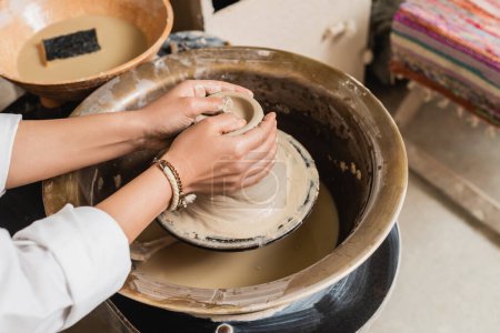 Cropped view of young craftswoman shaping wet clay on pottery wheel near blurred bowl with water and sponge in ceramic workshop, pottery studio workspace and craft concept