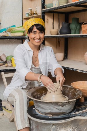 Fröhliche junge asiatische Künstlerin mit Kopftuch formt nassen Ton, während sie an der Töpferscheibe arbeitet und in Keramikwerkstatt, Töpferatelier und Bastelkonzept vor die Kamera blickt
