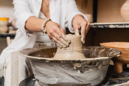 Vue recadrée d'une jeune femme artisan en vêtements de travail façonnant de l'argile humide tout en travaillant sur une roue de poterie près d'un bol à l'arrière-plan, un atelier de poterie et un concept artisanal