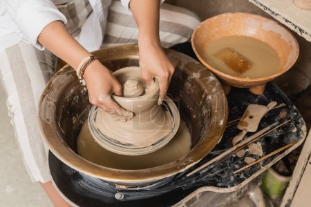 Top view of young female artisan molding wet clay on pottery wheel near tools and bowl with water in ceramic workshop, pottery studio workspace and craft concept