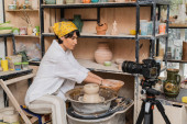 Asian female potter in workwear and headscarf looking at digital camera and working with bowl with water and wet clay on pottery wheel, pottery studio workspace and craft concept mug #663415126