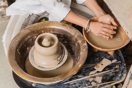 Vue du dessus de la jeune femme artisan en tenue de travail tenant éponge près du bol avec de l'eau et de l'argile humide sur roue de poterie tout en travaillant dans un atelier de céramique, atelier de poterie espace de travail et concept d'artisanat