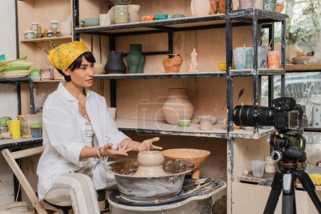 Asian female artisan in headscarf and workwear talking at digital camera on tripod while working with wet clay on pottery wheel near rack in art workshop, clay sculpting process concept