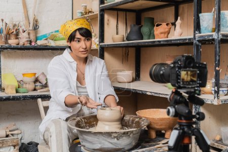 Asian female artisan in workwear and headscarf talking and gesturing at digital camera on tripod while working with clay on pottery wheel in art workshop, clay sculpting process concept