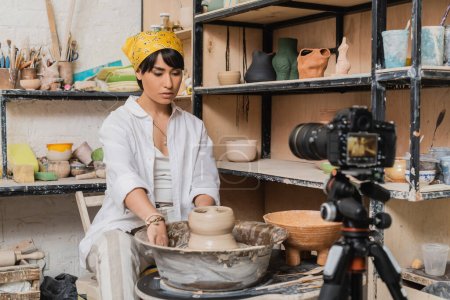 Young asian artist in headscarf and workwear working with wet clay on pottery wheel near blurred digital camera on tripod in ceramic workshop, clay sculpting process concept