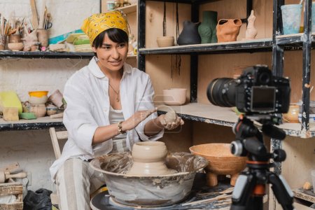 Positive asian female artist in headscarf holding wet clay and looking at digital camera on tripod near pottery wheel in art workshop, clay sculpting process concept