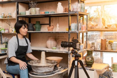 Photo for Young asian brunette female potter in apron looking at digital camera on tripod while sitting near pottery wheel and sculptures on rack in ceramic workshop, pottery tools and equipment - Royalty Free Image