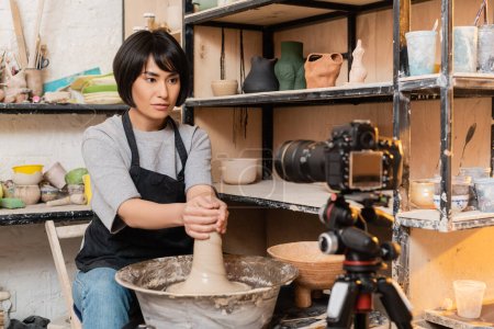 Young asian brunette craftswoman in apron molding wet clay on pottery wheel near blurred digital camera and sculptures on rack in workshop, pottery tools and equipment