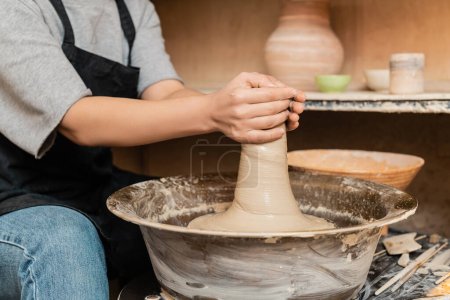 Téléchargez les photos : Vue recadrée d'une jeune artisan femelle dans un tablier moulant de l'argile humide sur une roue de poterie et travaillant près d'outils et d'un bol dans un atelier de céramique floue, des outils et des équipements de poterie - en image libre de droit