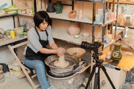 Junge asiatische Kunsthandwerkerin in Schürze beim Formen von nassem Ton auf Töpferscheibe in der Nähe von Schüssel mit Wasser und Werkzeug in der Nähe von Digitalkamera in der Keramikwerkstatt im Hintergrund, Töpferwerkzeuge und Ausrüstung