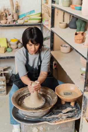 Joven mujer asiática artesana en delantal moldeado arcilla mojada en la rueda de cerámica cerca de cuenco con agua y herramientas mientras trabaja en taller de cerámica, herramientas de cerámica y equipo