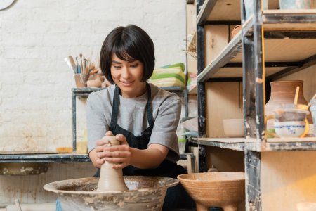 Sonriente joven artista femenina asiática en delantal moldeado de arcilla en la rueda de cerámica mientras trabaja cerca de rack y esculturas en taller de cerámica en el fondo, herramientas de cerámica y equipo