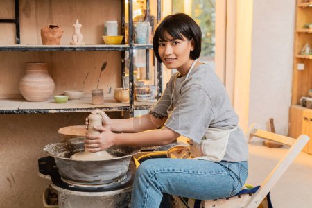 Fröhlicher und tätowierter junger asiatischer Kunsthandwerker in Schürze blickt in die Kamera, während er in der Keramikwerkstatt bei Sonnenuntergang feuchten Ton auf der Töpferscheibe formt, Töpferwerkzeuge und Geräte