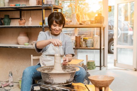 Lächelnde junge asiatische Künstlerin in Schürze gießt Wasser auf Ton auf Töpferscheibe in der Nähe von Werkzeugen und Schale in verschwommener Werkstatt bei Sonnenuntergang, Kunsthandwerker schafft einzigartige Keramikstücke