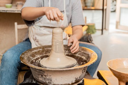 Ausgeschnittene Ansicht einer Kunsthandwerkerin in Schürze, die Wasser auf Ton gießt, während sie an der Töpferscheibe in einer verschwommenen Keramikwerkstatt im Hintergrund arbeitet, Kunsthandwerkerin, die einzigartige Keramikstücke schafft