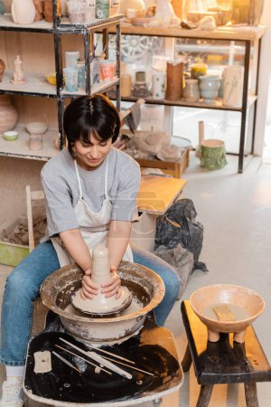 Jeune artisan asiatique brune en tablier moulant de l'argile humide sur une roue de poterie près d'un bol avec de l'eau et des outils dans un atelier de céramique floue au coucher du soleil, artisan créant des pièces de poterie uniques
