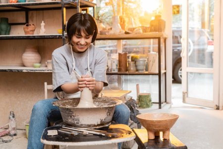 Téléchargez les photos : Joyeux jeune potier asiatique femelle en tablier moulant de l'argile sur la roue de poterie près du bol et des outils en atelier de céramique à l'arrière-plan au coucher du soleil, artisan créer des pièces de poterie uniques - en image libre de droit