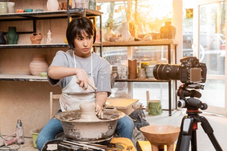 Jeune potier asiatique femelle en tablier pointant du doigt tout en travaillant avec de l'argile sur la roue de poterie et en regardant l'appareil photo numérique en atelier au coucher du soleil, artisan créer des pièces de poterie uniques