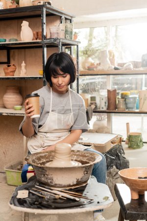 Junge asiatische Töpferin in Schürze mit Kaffee to go und Blick auf Ton auf Töpferscheibe in der Nähe von Werkzeugen und Schale in verschwommener Töpferei-Werkstatt, Kunsthandwerker schafft einzigartige Keramikstücke