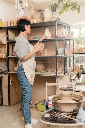 Joven mujer asiática artesana en delantal sosteniendo café para ir y escultura cerca de arcilla en rueda de cerámica y estante en taller de cerámica en el fondo, artesano creando piezas únicas de cerámica
