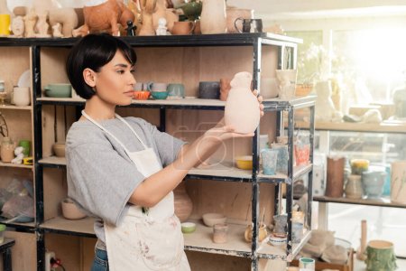 Jeune femme artisanale asiatique brune dans un tablier tenant une sculpture en argile près d'un rack avec des étagères dans un atelier en céramique floue à l'arrière-plan au coucher du soleil, artisan créant des pièces de poterie uniques