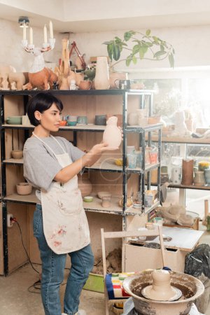 Junge brünette asiatische Künstlerin in Schürze, die Skulptur hält, während sie in der Nähe von Ton auf Töpferscheibe und Gestell im verschwommenen Keramik-Atelier bei Sonnenuntergang steht, Tonbildhauerei-Prozess-Konzept
